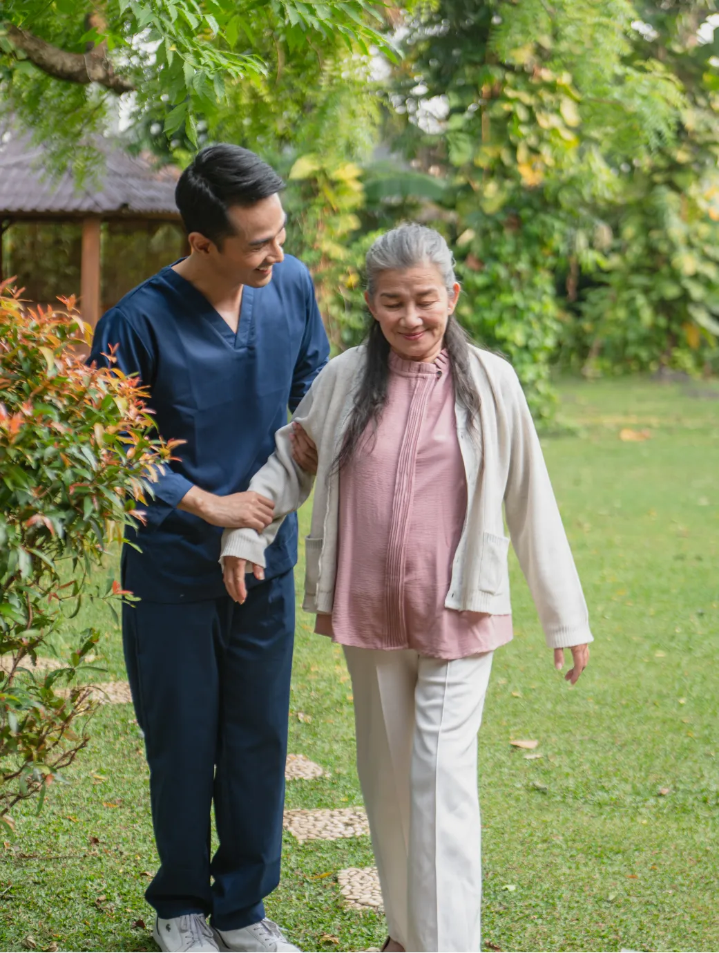 about-1 A caregiver in blue scrubs helps an elderly woman walk outside on a garden path.