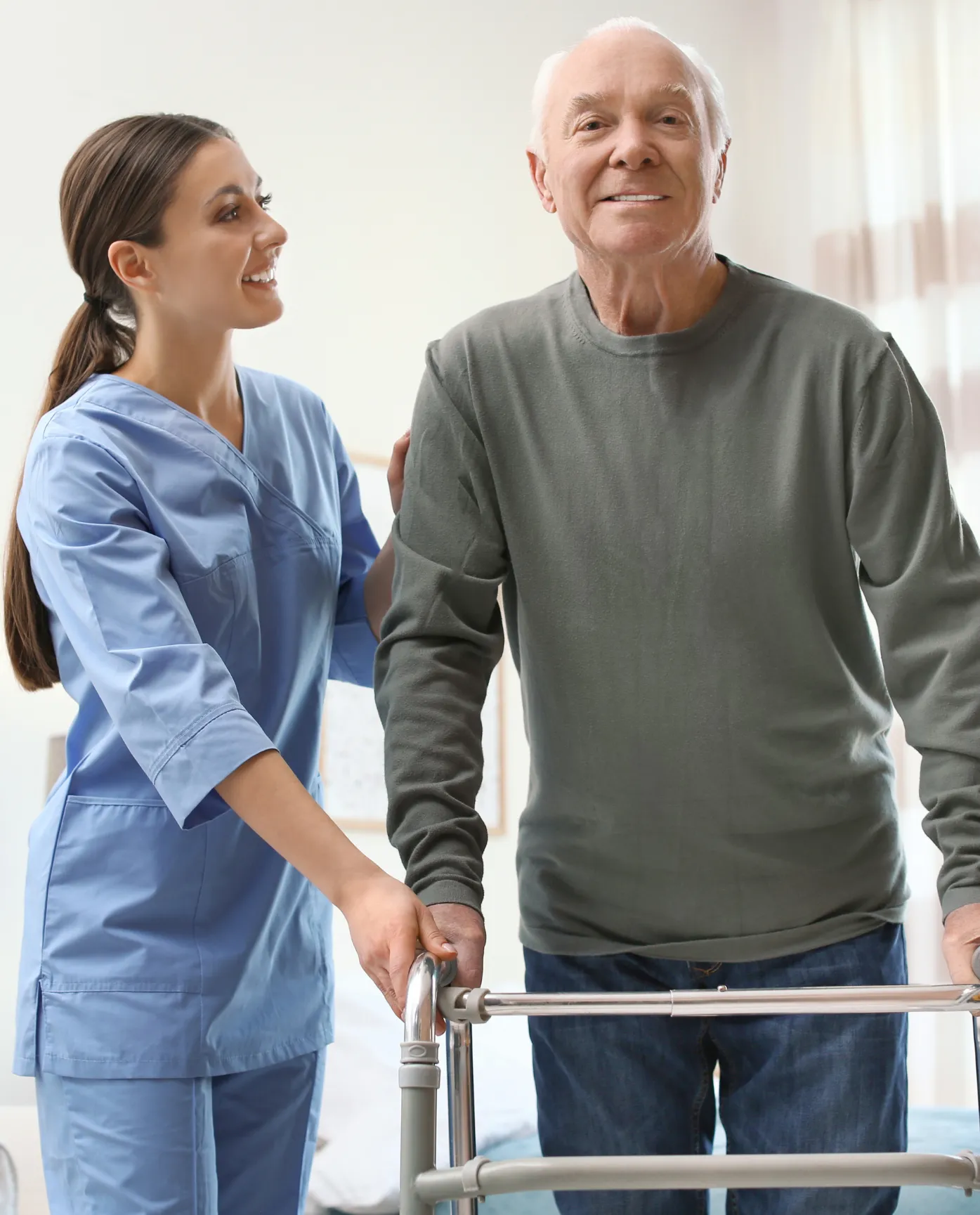 Young nurse helps elderly man with a walker, both smiling in a bright, comfortable room.