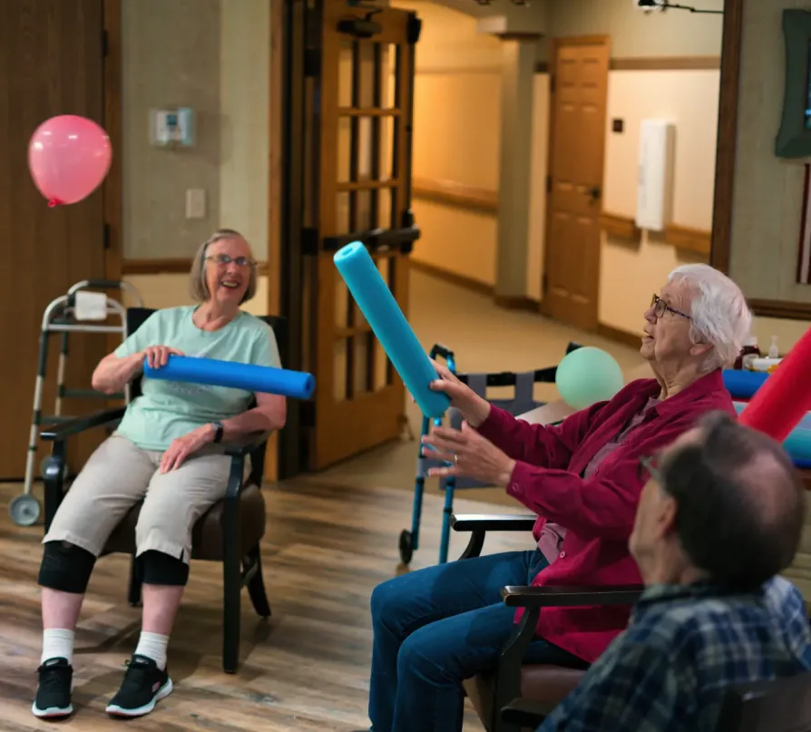 activities-1 Three seniors sitting and playing with pool noodles and balloons in a brightly lit room, smiling and laughing.