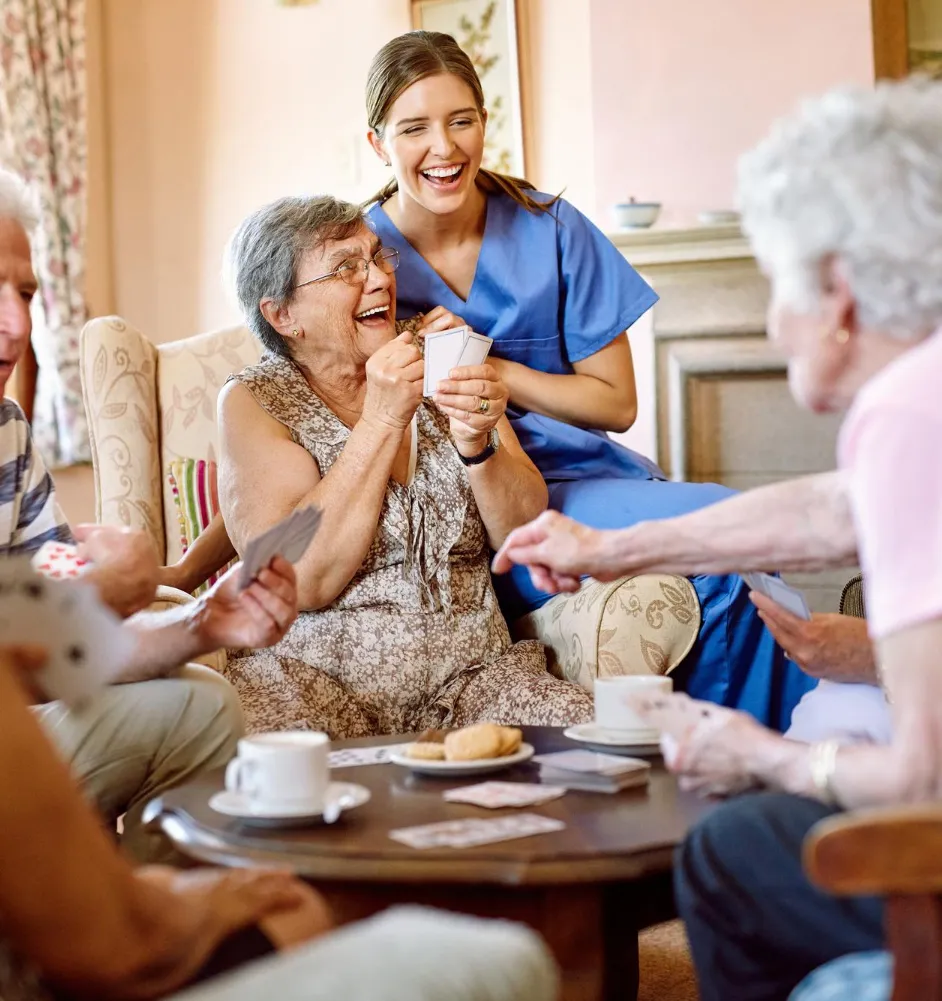 activities-w Elderly people and a caregiver laugh and play cards together around a coffee table with drinks and snacks.