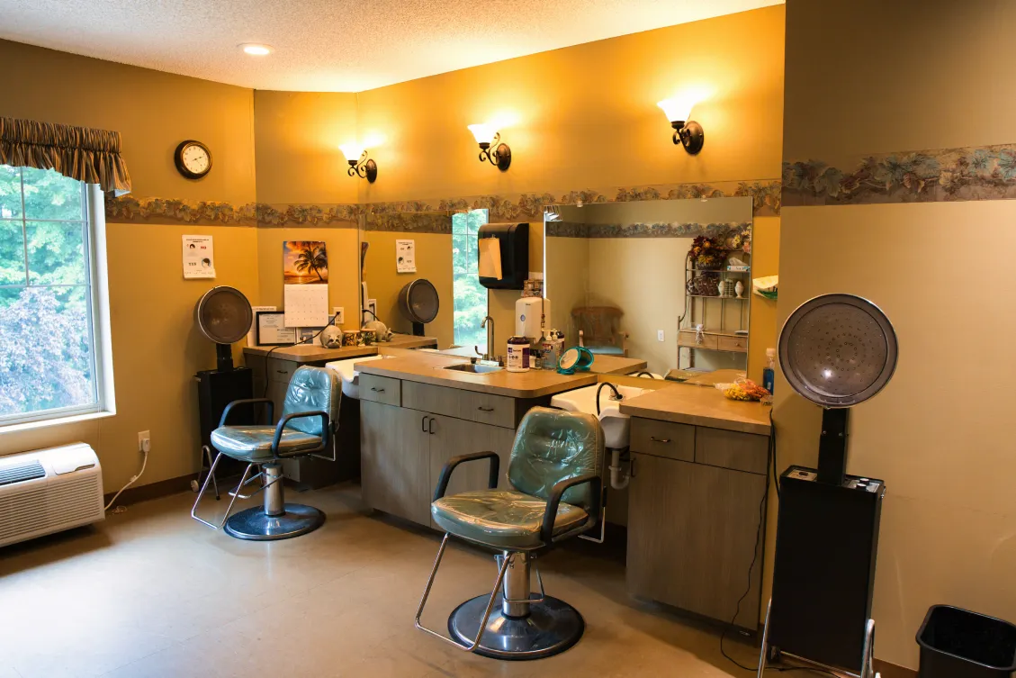 A salon with two chairs, mirrors, hair dryers, and warm yellow walls, lit by wall lamps.