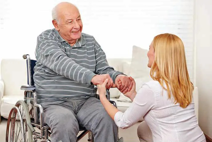 assisted-1 Smiling elderly man in a wheelchair holds hands with a woman kneeling beside him in a bright room.