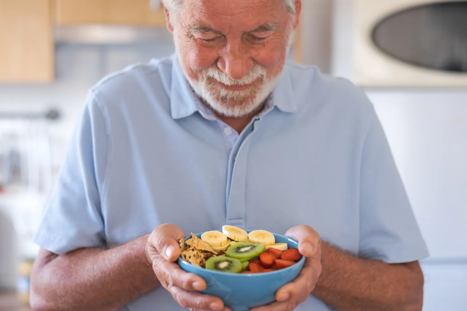 Smiling older man in a blue shirt holds a bowl of fruit, including banana, kiwi, and strawberries.