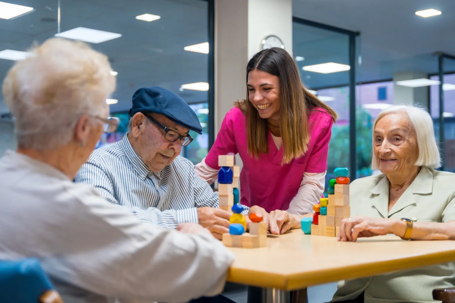 b3 A smiling caregiver helps three elderly people stack blocks at a table in a bright room.