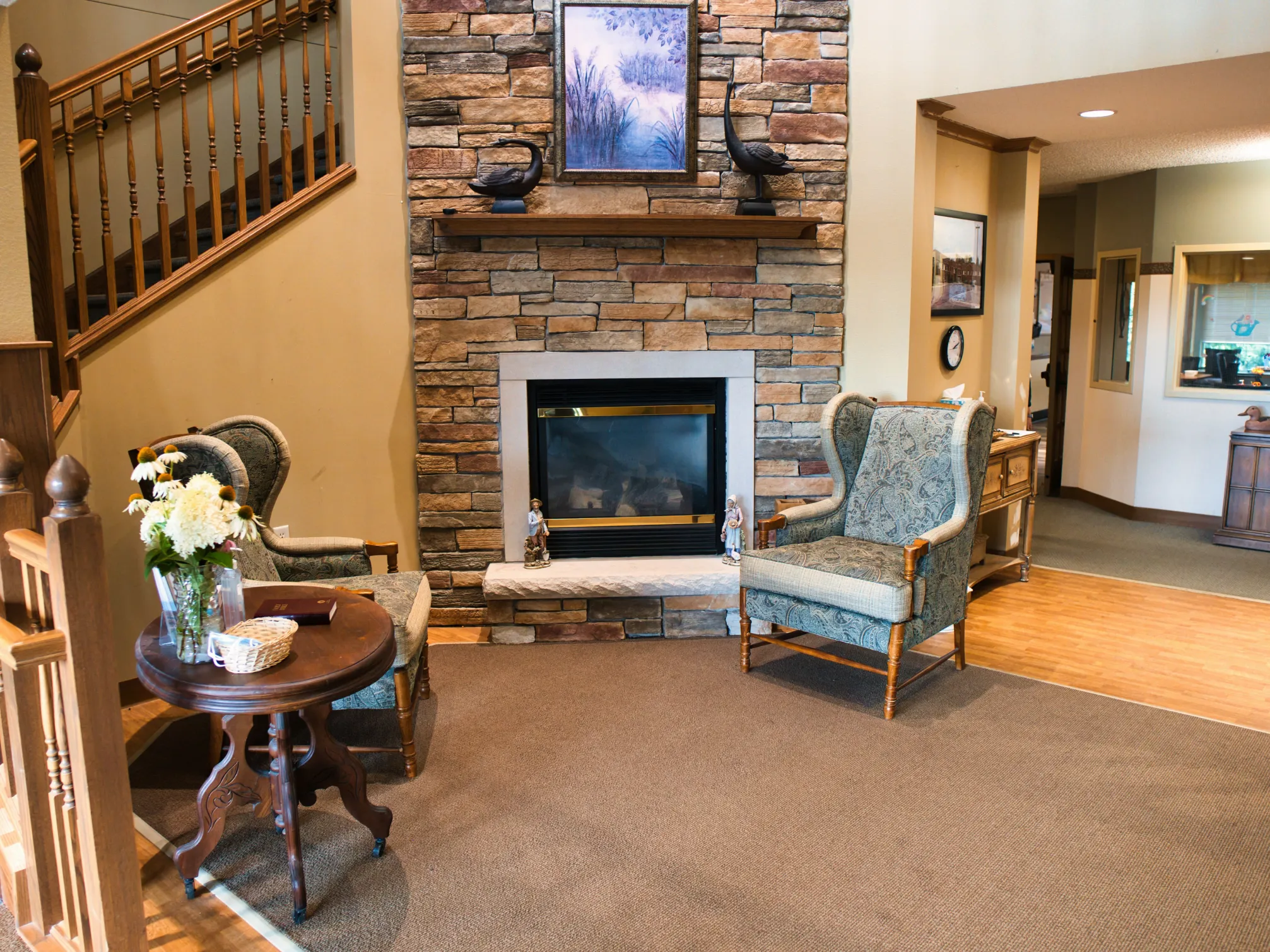 Two armchairs sit by a stone fireplace with a painting above and a table with flowers in between.