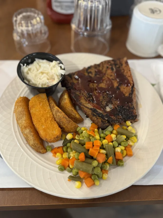 Plate with BBQ meat, potato wedges, coleslaw, and mixed vegetables on a table.