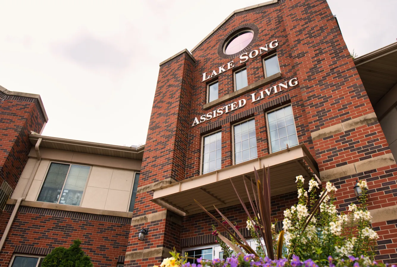 Red brick building with “Lake Song Assisted Living” sign, flowers in front, photographed from below.