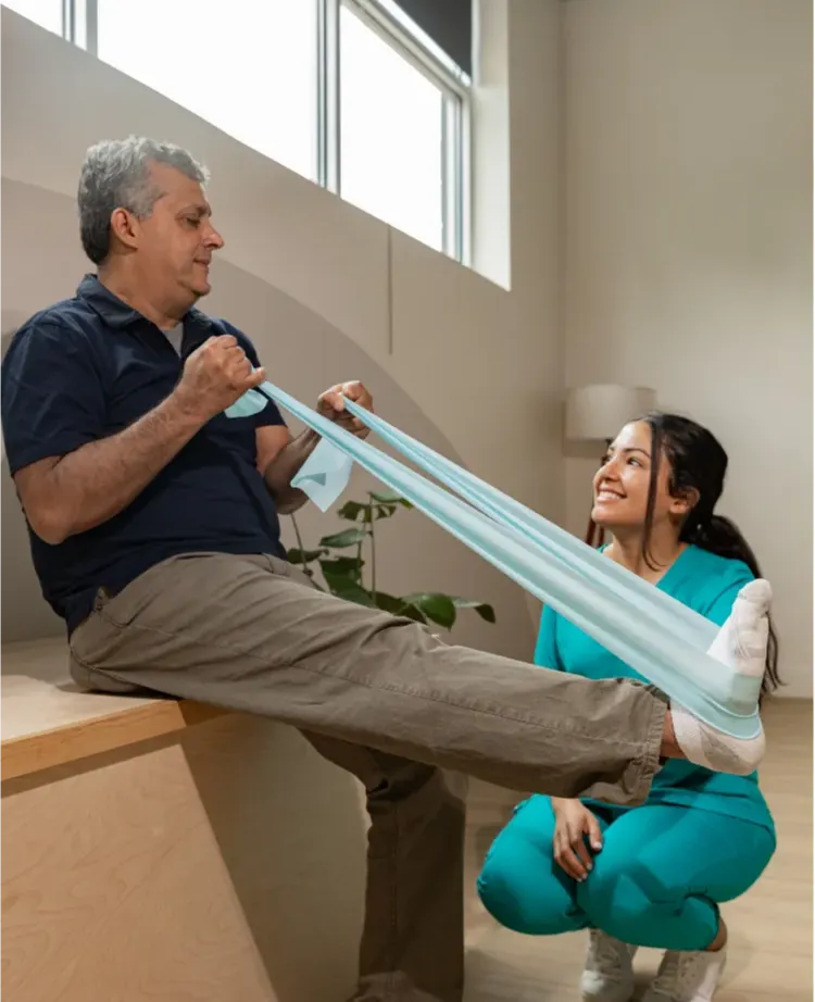 A man uses a resistance band for leg exercises with help from a smiling female physical therapist.