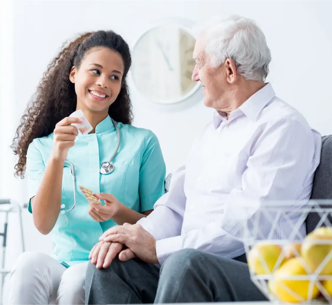 Smiling nurse gives medicine and water to an elderly man sitting on a couch in a bright room.