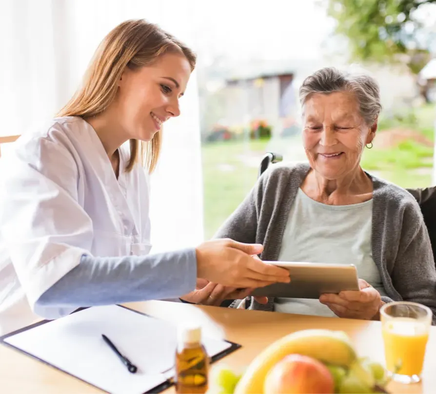 A caregiver shows an elderly woman a tablet at a table with fruit, both smiling and engaged.
