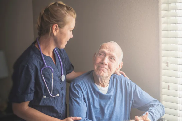 Nurse with stethoscope comforts an elderly man sitting by a window with gentle sunlight coming through blinds.