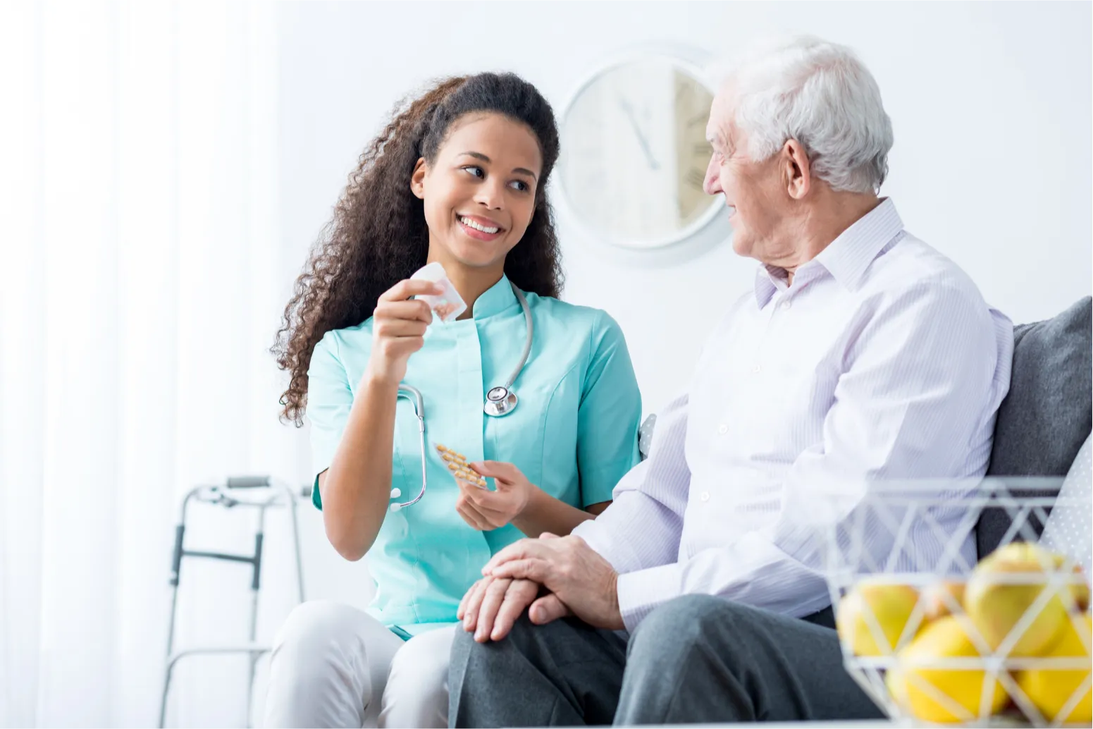 A nurse hands medication to an elderly man sitting on a couch, both smiling at each other.