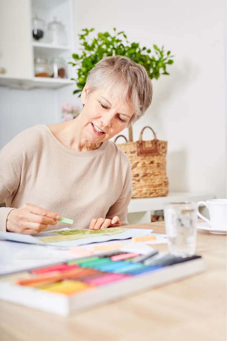 Smiling older woman drawing with colored pencils at a table, with a cup and art supplies nearby.