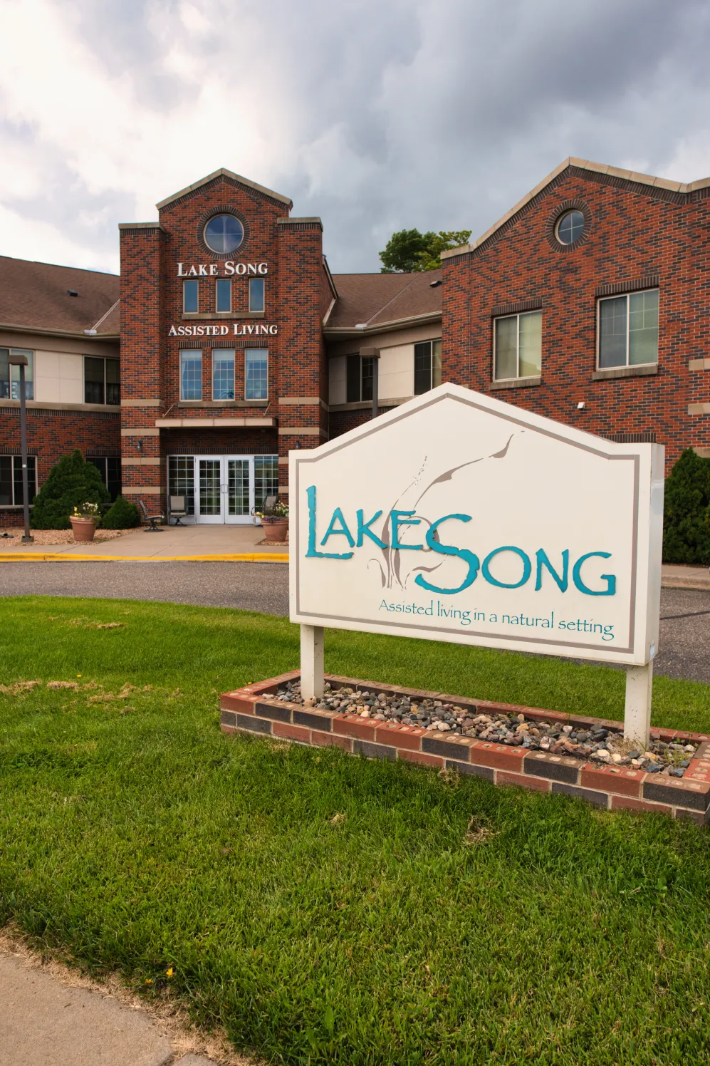 our-story Brick building with “LakeSong Assisted Living” sign and green lawn in front, under a cloudy sky.