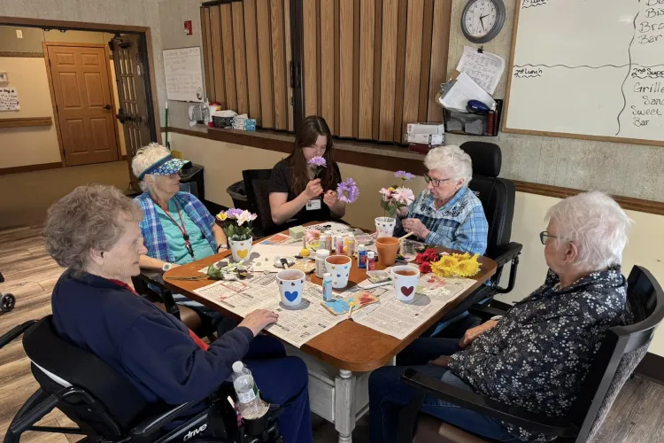 Five women sit around a table doing arts and crafts with flowers and paint in a community room.