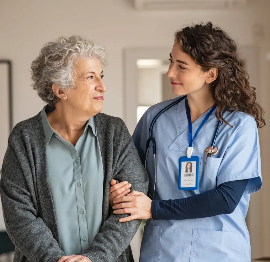 Young nurse smiles and supports an older woman, both looking at each other warmly in a bright room.